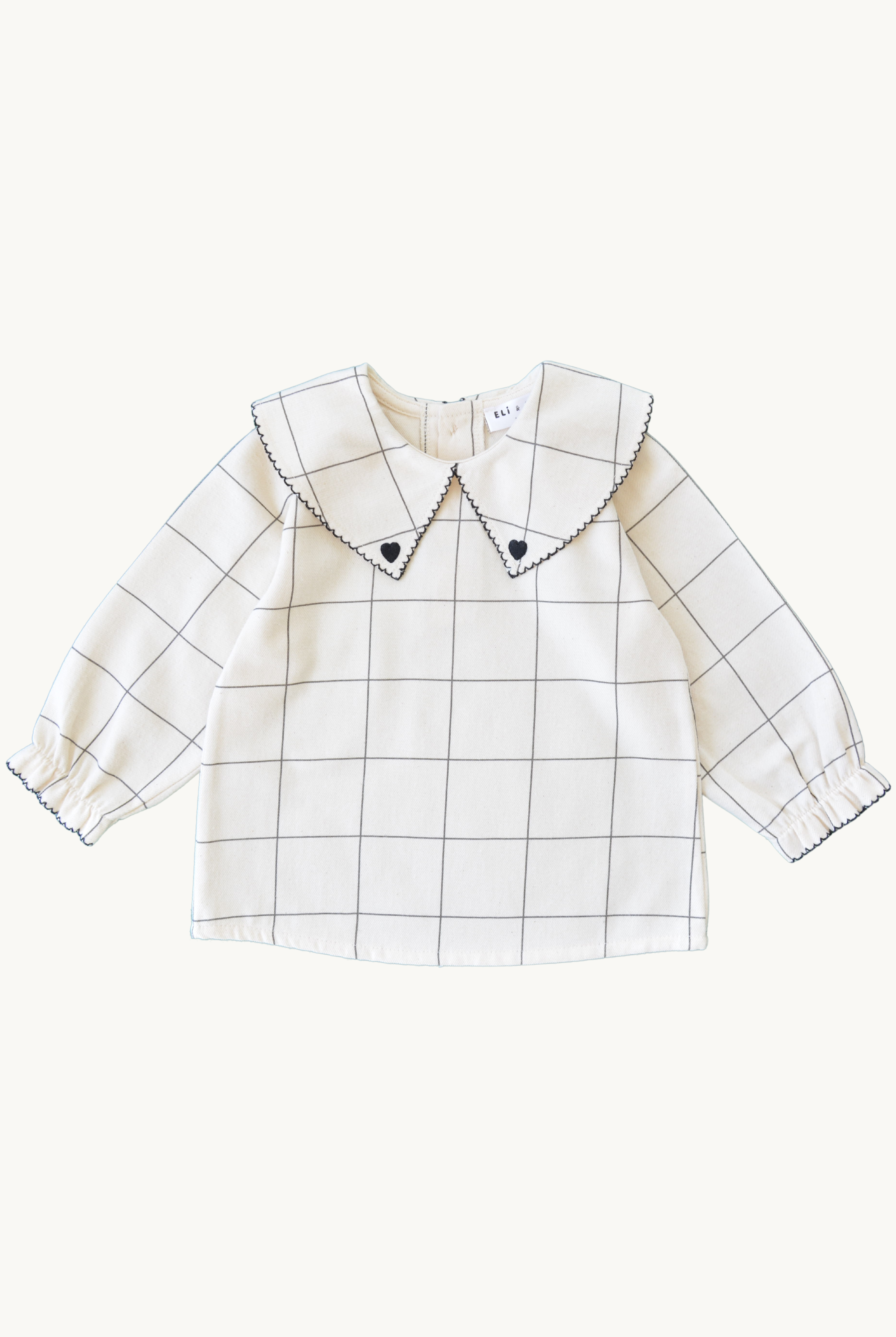 Beige blouse with black trim detailed collar with 'heart' embroidery, black trim on elastic cuffed sleeves, and coconut buttons on the back, displayed against a white background.